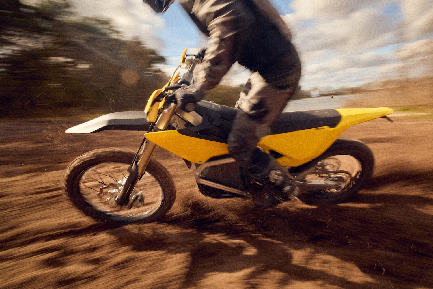 Motorcyclist riding a yellow dirt bike rapidly on a dirt track, kicking up dust.