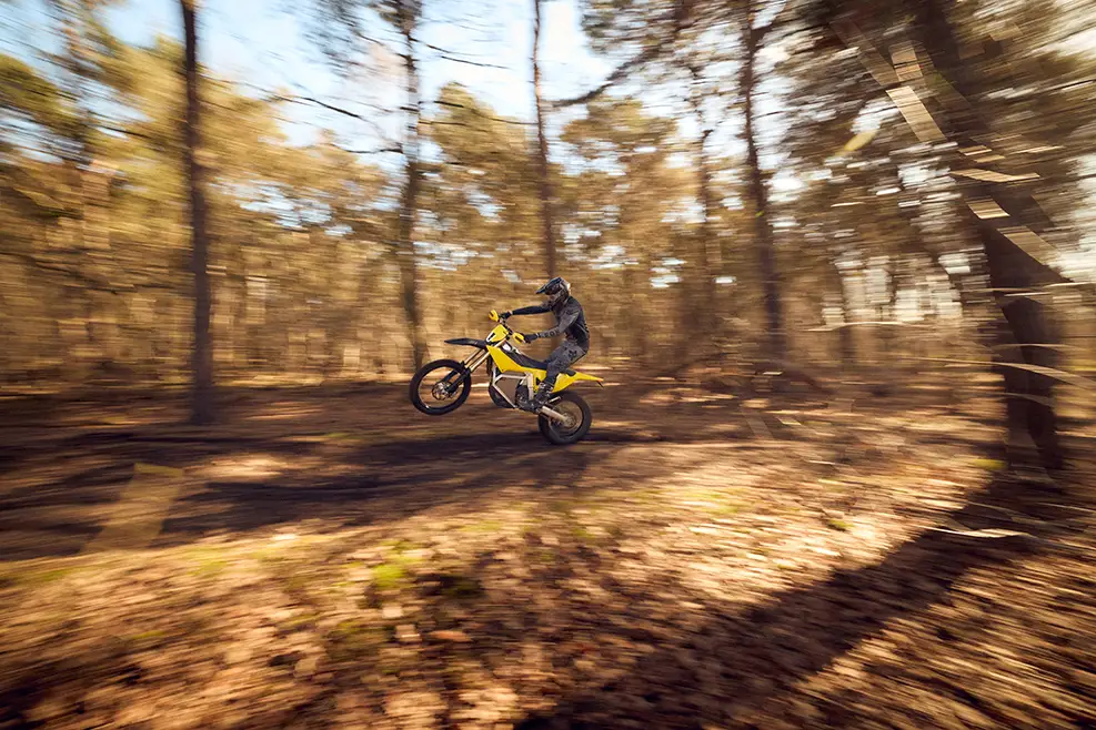 Rider performing a wheelie on a yellow dirt bike in a sunlit forest.