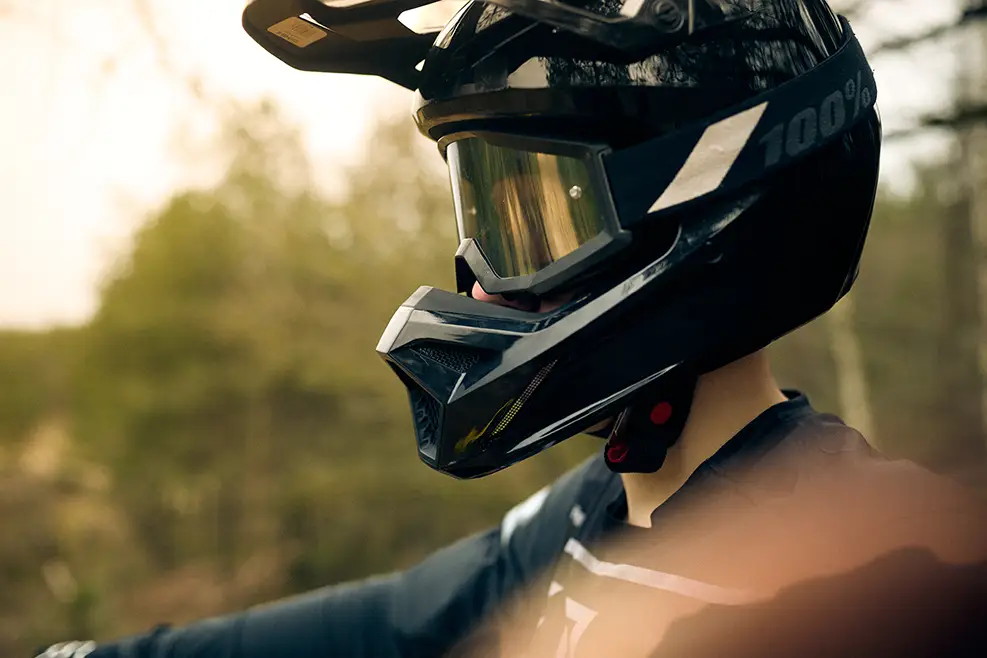 Person wearing a black full-face helmet with goggles outdoors in a blurred natural setting.