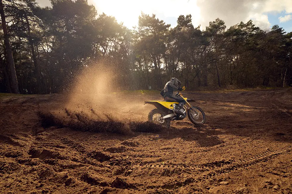 Person riding a yellow dirt bike kicking up a large spray of dirt on a forest trail at sunset.