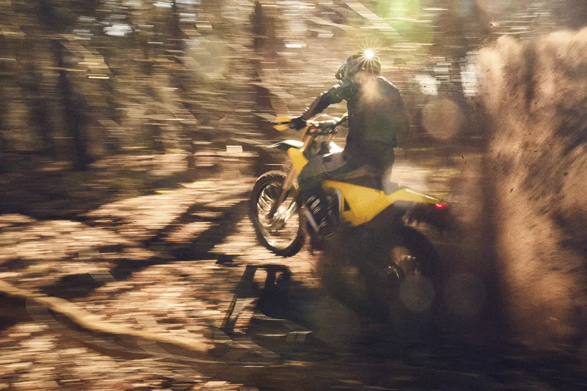 Person riding a yellow dirt bike through a forest kicking up dust at high speed.